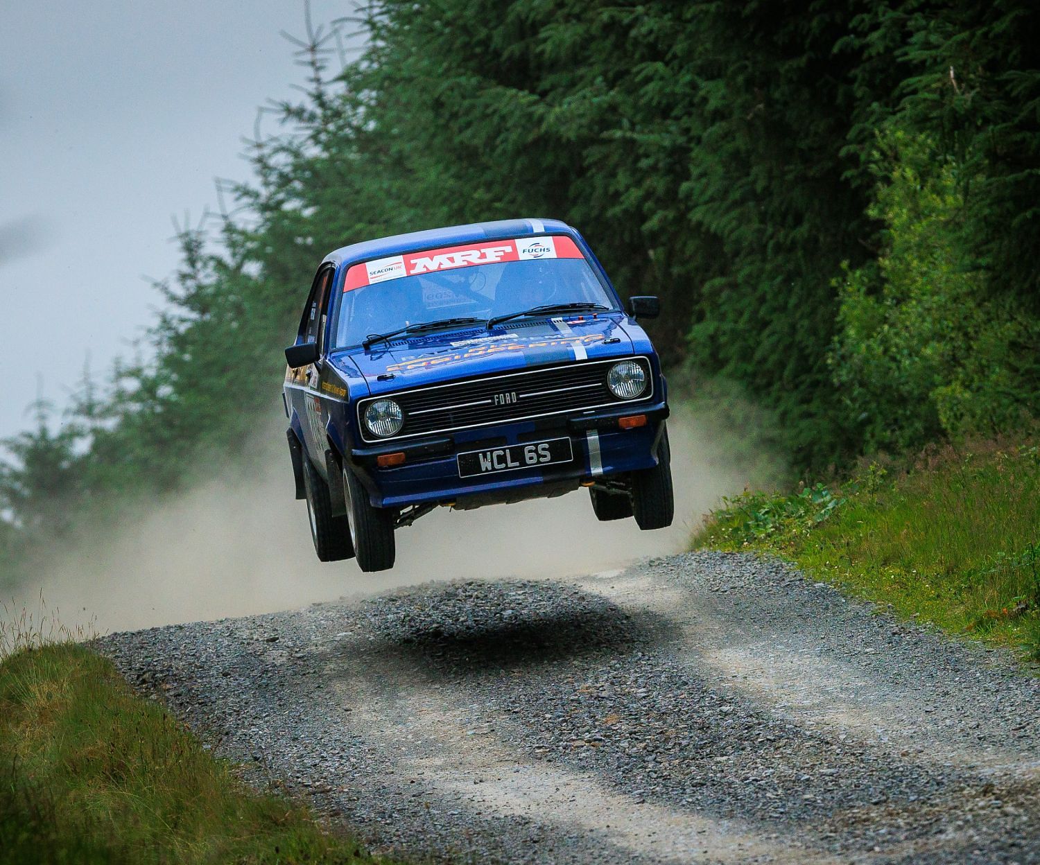 Blue Ford Escort MK2 airborne over a forest crest