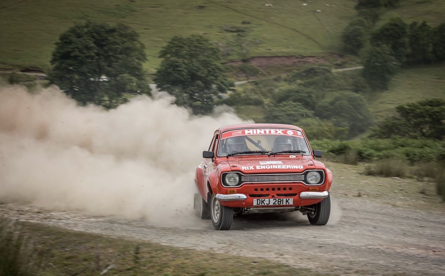 Red Ford Escort rally car kicking up dust on a gravel stage